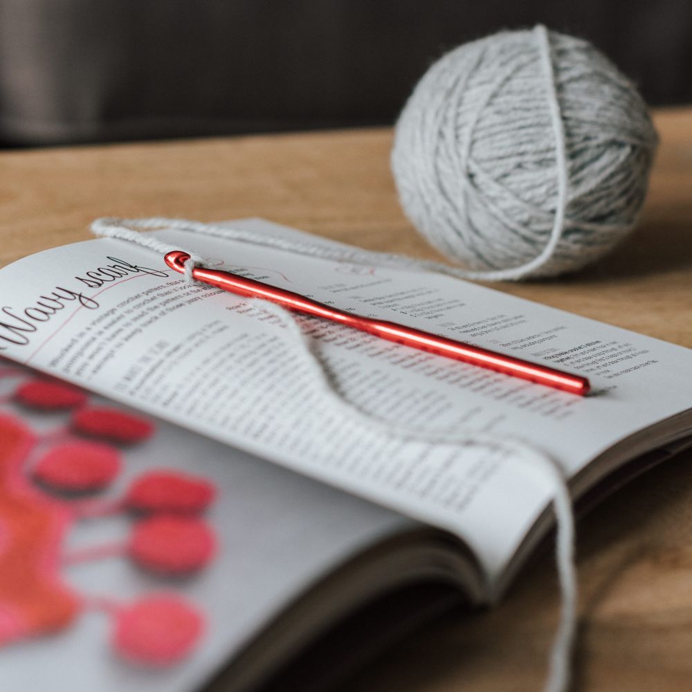A crochet pattern magazine open on a light wooden table with a crochet hook and yarn